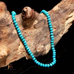 May include: A turquoise gemstone necklace composed of small, round beads. The necklace is displayed on a piece of weathered wood, with a dark background. The beads are a vibrant, consistent blue-green color.