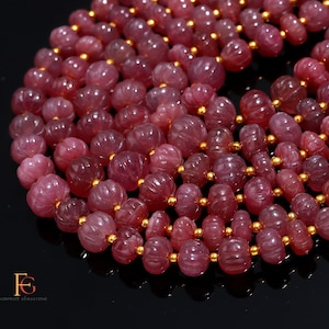 May include: Close-up of a necklace featuring carved, round, pink gemstones with gold-colored spacers. The gemstones have a ribbed texture and are strung together. The background is black.