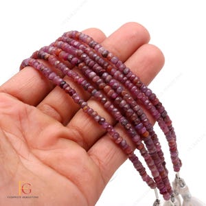 May include: A strand of pink ruby beads, faceted and round, strung on a white cord. The beads are approximately 3mm in diameter and are arranged in a straight line. The beads are displayed on a hand.