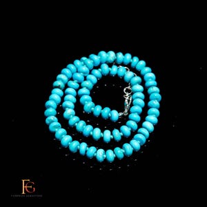 May include: A turquoise beaded necklace with a silver clasp. The necklace is composed of round, light blue beads. The beads are strung together to form a necklace. The necklace is displayed on a black surface.