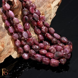 May include: A strand of deep red ruby gemstone beads, each bead is a smooth, oval shape. The beads are arranged in a loose, flowing pattern, creating a visually appealing necklace.