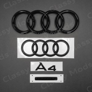 Op de afbeelding: Zwart Audi-embleem met vier in elkaar grijpende ringen, een zwart A4-embleem en een zwart rechthoekig embleem met een zilveren balk. De emblemen bevinden zich op een witte achtergrond.
