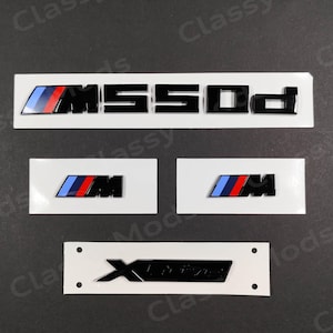 Könnte beinhalten: Ein Satz von drei schwarzen und blauen Autoemblemen. Das erste Emblem ist ein schwarzes "M" mit blauen und roten Streifen. Das zweite Emblem ist ein schwarzes "M" mit blauen und roten Streifen. Das dritte Emblem ist ein schwarzes "X" mit einem schwarzen "Drive"-Text.