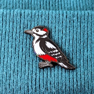 A black, white, and red woodpecker enamel pin with a rainbow tail feather detail, pinned to a teal knit beanie.
