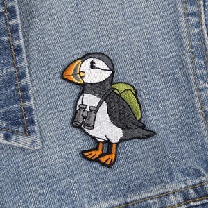 Può includere: Toppa ricamata raffigurante un pulcinella di mare dei cartoni animati con uno zaino e un binocolo. Il pulcinella di mare ha un corpo nero, bianco e grigio con un becco e zampe arancioni. La toppa è cucita su una giacca di jeans blu.