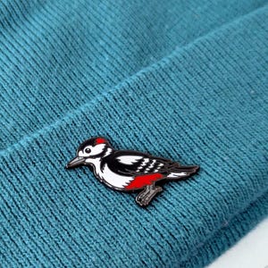 A black, white, and red woodpecker enamel pin, with its head slightly lowered and body angled, pinned to a teal knit beanie.