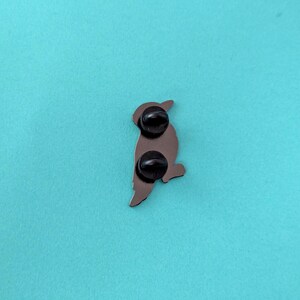 The back of a black woodpecker enamel pin, showing two black pin posts, against a teal background.
