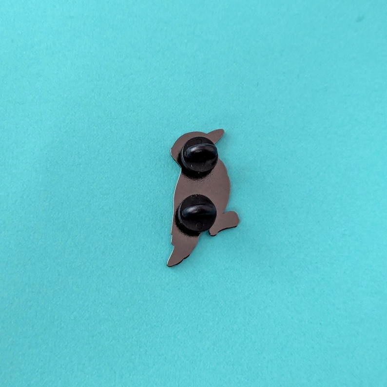 The back of a black woodpecker enamel pin, showing two black pin posts, against a teal background.