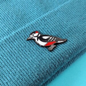A black, white, and red woodpecker enamel pin, with its head slightly lowered, pinned to a teal knit beanie.