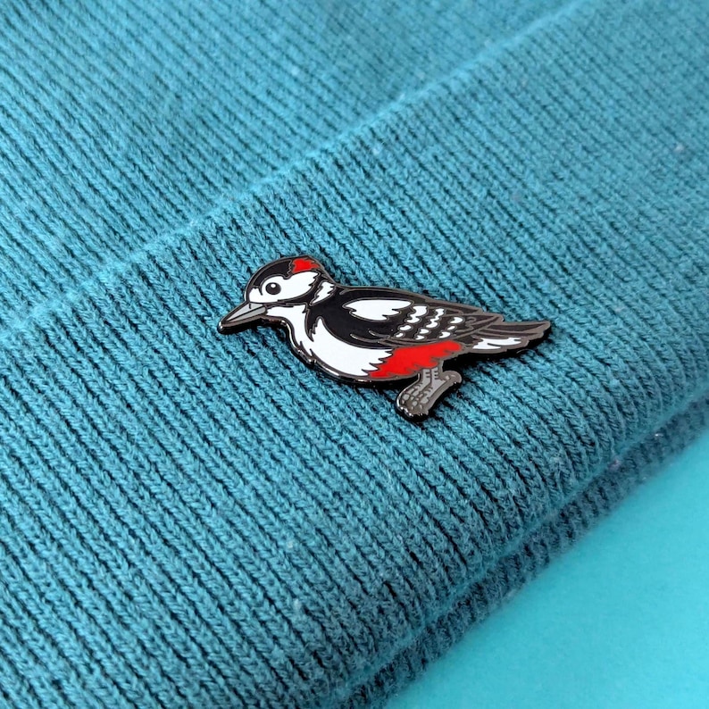 A black, white, and red woodpecker enamel pin, with its head slightly lowered, pinned to a teal knit beanie.