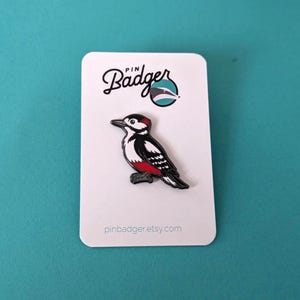 A black, white, and red woodpecker enamel pin on a white backing card with "PIN Badger" logo, on a light blue background.