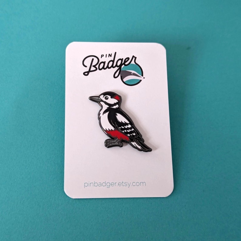 A black, white, and red woodpecker enamel pin on a white backing card with "PIN Badger" logo, on a light blue background.