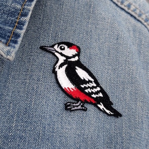 May include: An embroidered patch featuring a woodpecker with black, white, and red thread detailing. The bird is depicted in profile, showcasing a white chest, black wings with white markings, a red patch on the head, and a red patch on the belly.