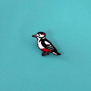 A black, white, and red woodpecker enamel pin, facing left, with a red patch on its head and under its tail, against a teal background.