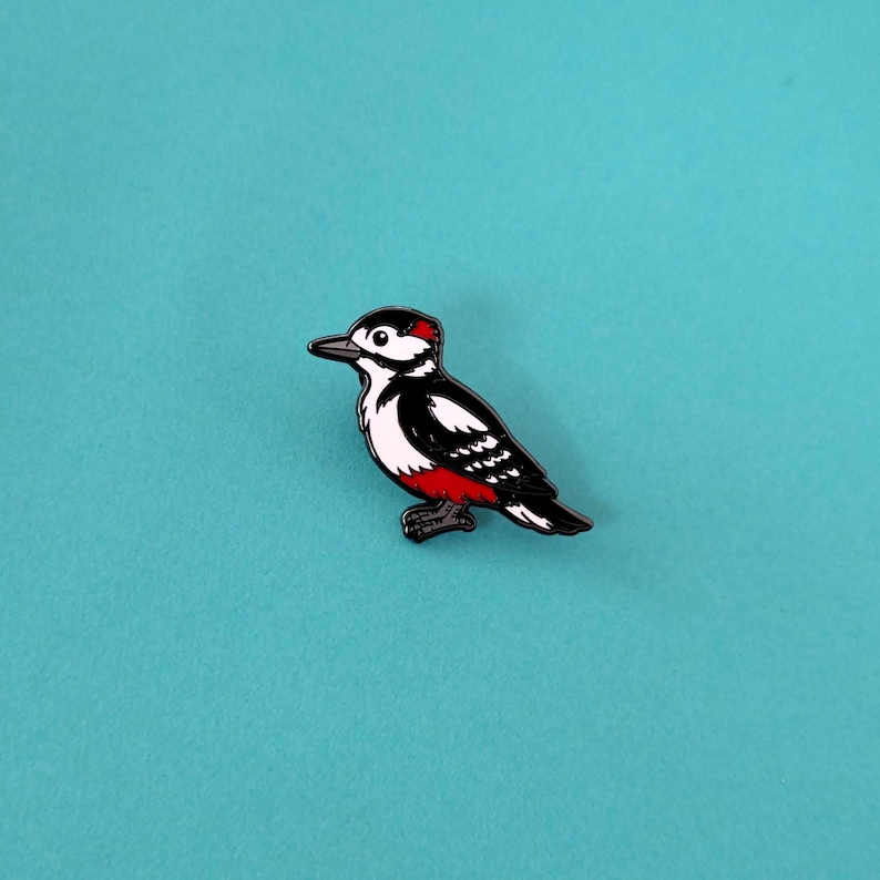 A black, white, and red woodpecker enamel pin, facing left, with a red patch on its head and under its tail, against a teal background.