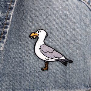 Pode incluir: Um emblema bordado de uma gaivota branca e cinzenta com bico e patas amarelas, segurando um pequeno objeto amarelo no bico, costurado num tecido de ganga azul claro com trama diagonal visível e uma costura no lado esquerdo.