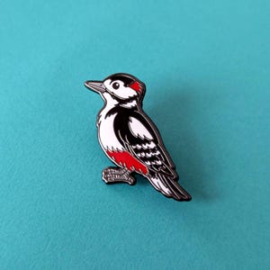 A black, white, and red woodpecker enamel pin, facing left, on a light blue background.
