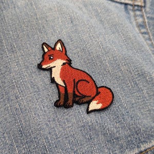 May include: An embroidered patch featuring a sitting fox with rust-orange fur, a white chest, and a white-tipped tail. The fox has dark brown paws and ears with black outlines, positioned against a textured light blue denim fabric background.