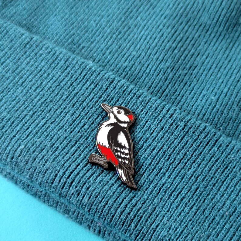A black, white, and red woodpecker enamel pin, with its head slightly raised, pinned to a teal knit beanie.