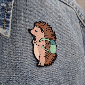 May include: An embroidered patch featuring a light pink hedgehog with brown quills, standing in profile and wearing a two-tone light green backpack. The patch is stitched onto a blue denim fabric background with visible seams and a textured weave.