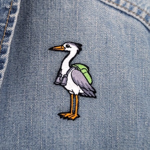 May include: An embroidered patch featuring a white and gray bird with an orange beak and feet. The bird is wearing a green backpack and has a pair of black binoculars hanging around its neck. The patch is attached to a blue denim fabric surface.