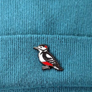 A black, white, and red woodpecker enamel pin, with its head facing forward, pinned to a teal knit beanie.