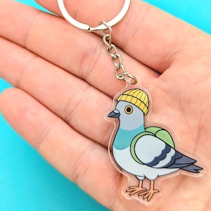 Può includere: Un portachiavi in acrilico trasparente con un piccione dei cartoni animati con un berretto giallo e uno zaino verde. Il piccione ha piume blu, grigie e bianche. Il portachiavi ha un anello e una catena in metallo argentato.