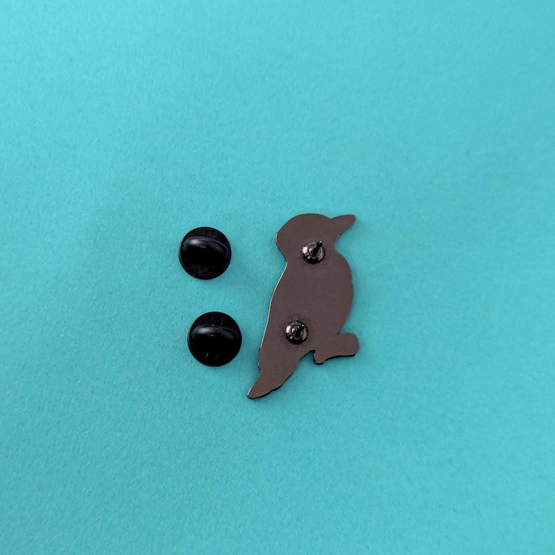 The back of a woodpecker enamel pin, showing two black pin posts, on a light blue background.