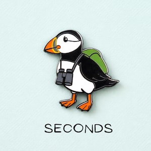 Può includere: Una spilla smaltata colorata raffigurante un pulcinella di mare dei cartoni animati con uno zaino verde e un binocolo. Il pulcinella ha un corpo bianco e nero, zampe e becco arancioni, su sfondo azzurro chiaro. La parola "SECONDS" è stampata sotto.