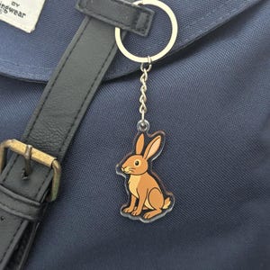 Puede incluir: Un llavero de conejo marrón y beige con una cadena y un anillo plateados. El llavero cuelga de una mochila azul marino. El conejo está sentado con las orejas erguidas.