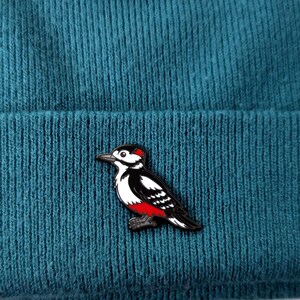 A black, white, and red woodpecker enamel pin, with its head facing forward, pinned to a teal knit beanie.