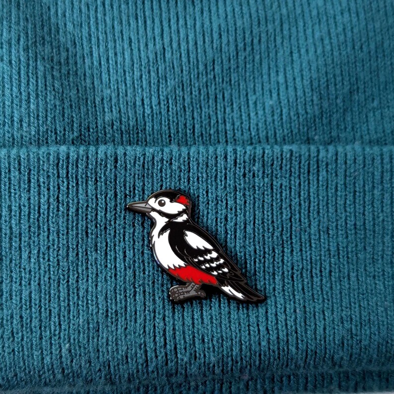 A black, white, and red woodpecker enamel pin, with its head facing forward, pinned to a teal knit beanie.
