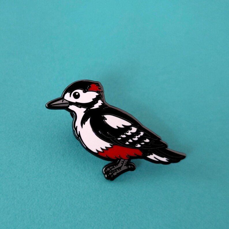 A black, white, and red woodpecker enamel pin, facing left, on a light blue background.