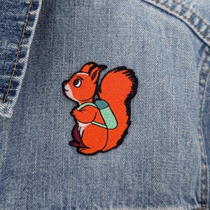 Peut inclure: Un écusson brodé représentant un écureuil orange vif avec une grande queue touffue et un sac à dos vert clair, fixé sur un tissu en jean bleu. L'écureuil est représenté de profil, tourné vers la gauche avec un poitrail blanc et des contours noirs.