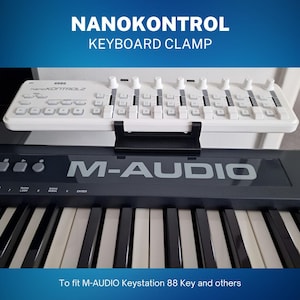 Könnte beinhalten: Eine weiße Tastaturklemme für einen Korg NanoKontrol2 MIDI-Controller. Die Klemme ist an einem schwarzen M-AUDIO Keystation 88 Key Keyboard befestigt. Der Text "To fit M-AUDIO Keystation 88 Key and others" ist am unteren Rand des Bildes sichtbar.