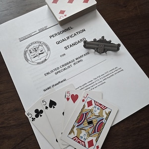 May include: A deck of playing cards with the five of diamonds visible on top, along with four other cards fanned out. A document titled "Personnel Qualification Standard" and a military pin are also present.