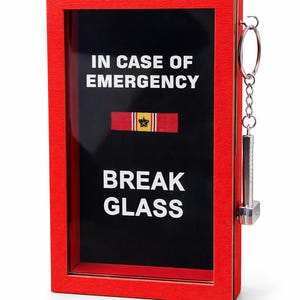Puede incluir: Una caja roja con el texto "IN CASE OF EMERGENCY BREAK GLASS" sobre un fondo negro. Un pequeño martillo de metal está sujeto al lateral con una cadena. Una cinta roja y dorada se muestra en el centro.