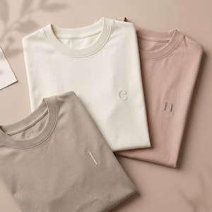 Könnte beinhalten: Drei Rundhals-T-Shirts in Creme, Taupe und Rosé. Jedes Shirt hat einen einzelnen gestickten Buchstaben oder eine Zahl: „e“, „1“ und „n“. Die Shirts sind ordentlich gefaltet und zeigen das minimalistische Design und die neutrale Farbpalette.
