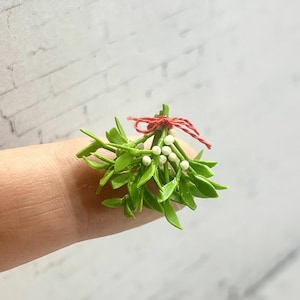 以下が含まれることがあります： 赤い紐で結ばれた、白い実のついた緑色のヤドリギの小さな枝。葉は鮮やかな緑色で、実は小さくて丸いです。お祝いのミニチュアホリデーデコレーション。