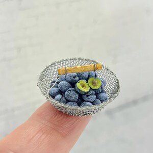 以下が含まれることがあります： 小さな濃い青色の丸い果物が入ったミニチュアのワイヤーメッシュバスケット。バスケットの上部には細い明るい茶色の木製ハンドルが付いています。果物が一つ半分にカットされており、鮮やかな緑色の果肉と小さな茶色の種が見えています。