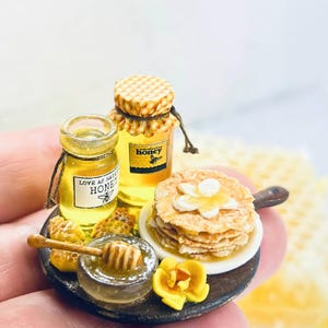 Könnte beinhalten: Eine Miniaturdarstellung auf einem dunklen runden Tablett mit zwei Honiggläsern mit Etiketten, auf denen Love As Sweet Honey und honey steht, einem Stapel kleiner Pfannkuchen auf einem weißen Teller, einem Honiglöffel und gelben Blumen.