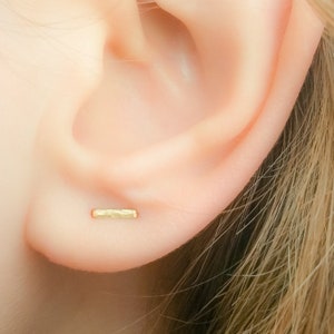 May include: A close-up of a person's ear with a small, gold, hammered bar earring. The earring is simple and minimalist.