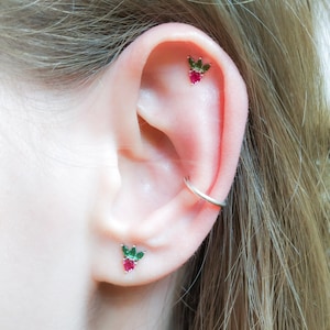 May include: Close-up of a person's ear with two small strawberry-shaped earrings with green leaves and a silver hoop earring in the upper cartilage.