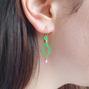 May include: Green and gold hoop earrings with a pink bead dangling from the bottom. The earrings are made of wire and have a simple, modern design.