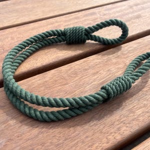 May include: A dark green rope with a loop at each end, knotted in the middle. The rope is made of twisted fibers and lies on a wooden surface. The rope is a decorative item.