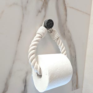 May include: A white rope toilet paper holder with a black accent piece. The holder is mounted on a white marble wall and holds a roll of white toilet paper. The rope is twisted and forms a loop.