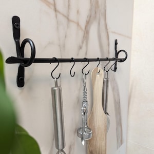 Op de afbeelding: Een zwarte metalen wandstang met decoratieve krulvormige uiteinden en zes bijpassende S-haken. De stang is met schroeven bevestigd en houdt diverse keukengerei vast, waaronder een metalen deegroller, een kurkentrekker en een houten spatel.