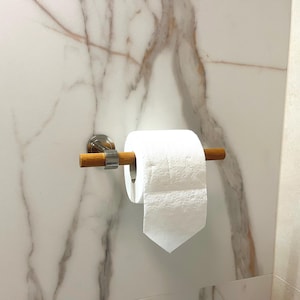 May include: A modern toilet paper holder with a wooden dowel and a brushed nickel finish. The holder is mounted on a white marble-patterned wall, with a roll of white toilet paper in place.
