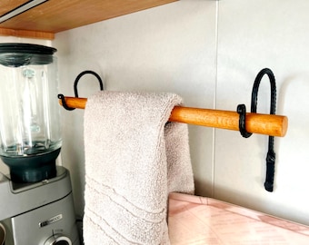 May include: A wooden towel rail with black metal fixings holds a folded beige towel. The rail is mounted on a white wall. A blender is visible in the background.