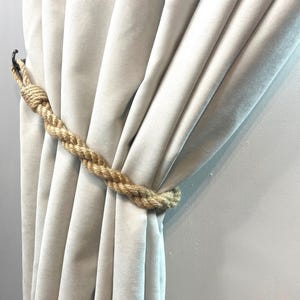 Puede incluir: Una cortina beige con un amarre de cuerda. La cuerda está hecha de fibras naturales y tiene un diseño de nudo simple.